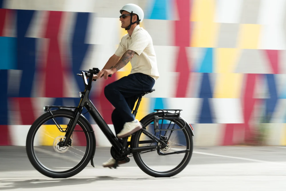 Homme à vélo électrique devant un mur coloré, portant un casque et des lunettes de soleil.